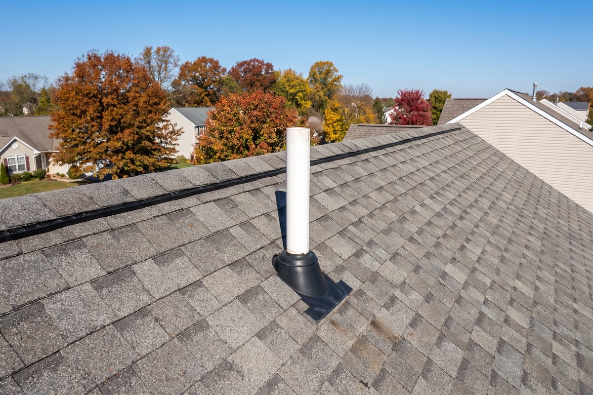 White PVC radon vent pipe rising above the asphalt shingle roof of a newer two-story house in York County, Pennsylvania, with a rubber flashing boot at its base and autumn trees behind