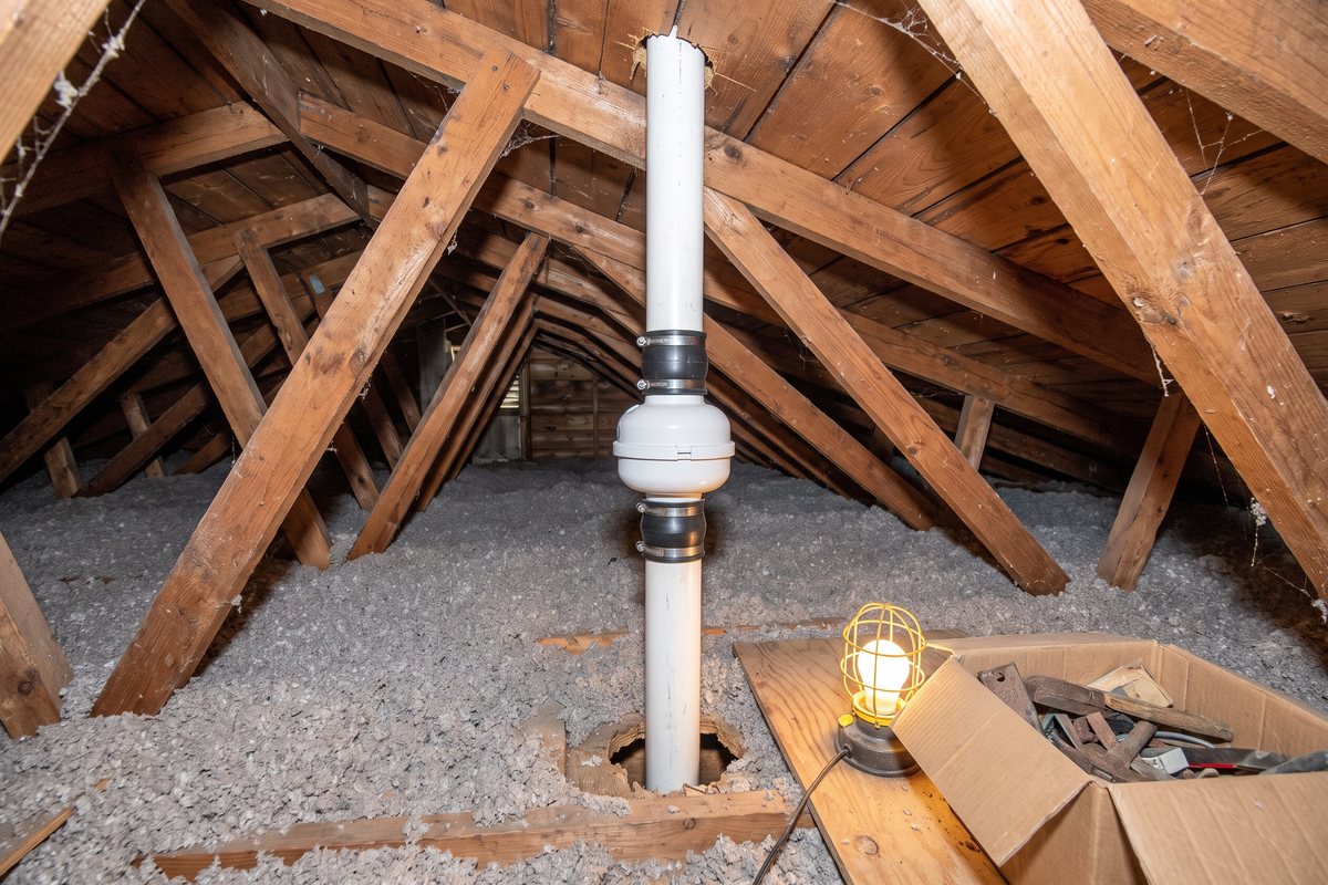 Radon fan mounted vertically on a white PVC vent pipe in an unfinished Pennsylvania attic, between wooden rafters above blown-in insulation, with the pipe continuing up through the roof deck